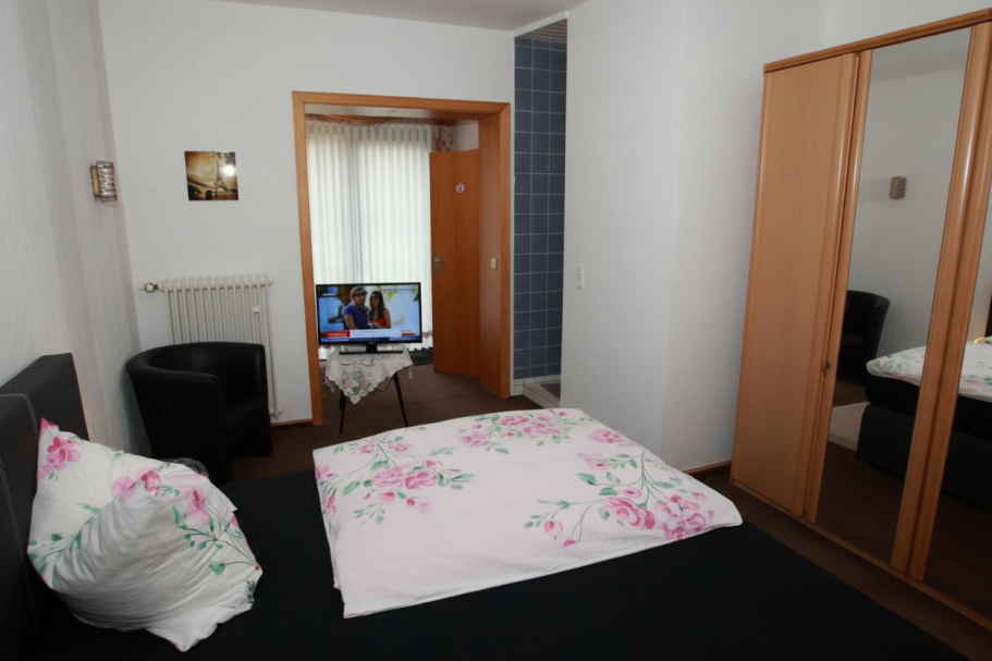 Helles Zimmer mit Bett, Fernseher, Schrank und Sitzbereich. Blütenmuster auf der Bettdecke.